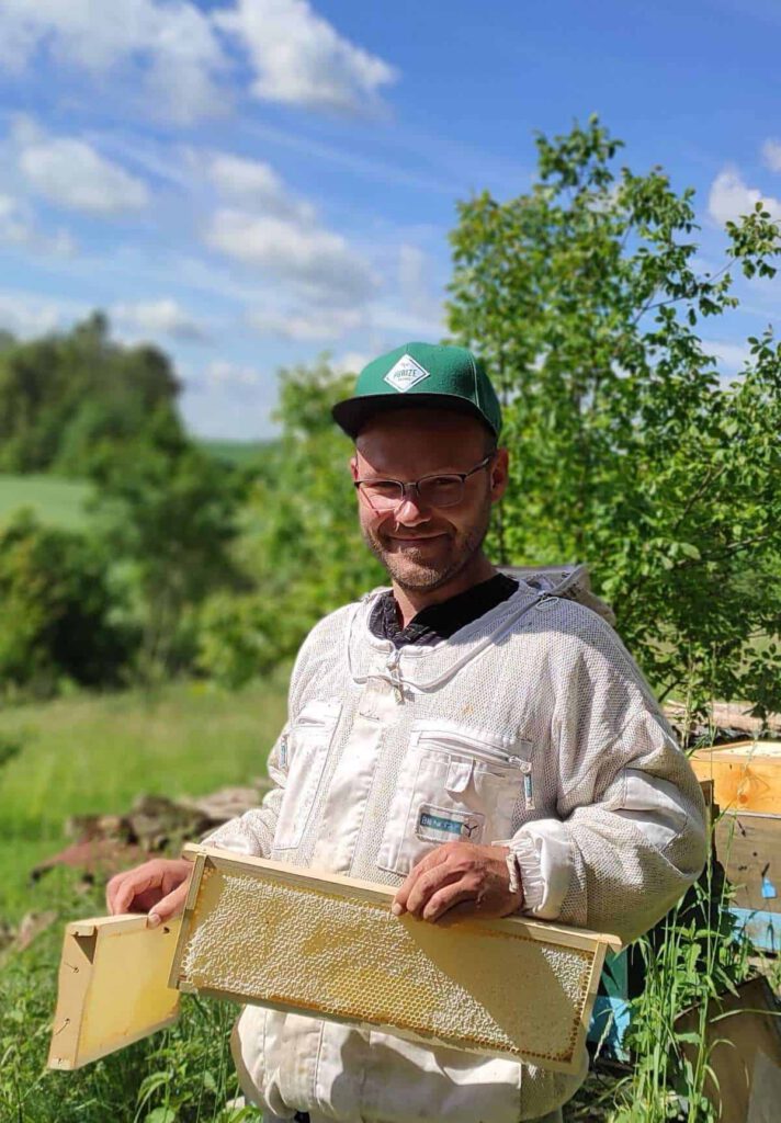 Florian in seiner Imker-Kleidung. Er steht auf der Wiese und hält zwei Bienenwaben in der Hand.