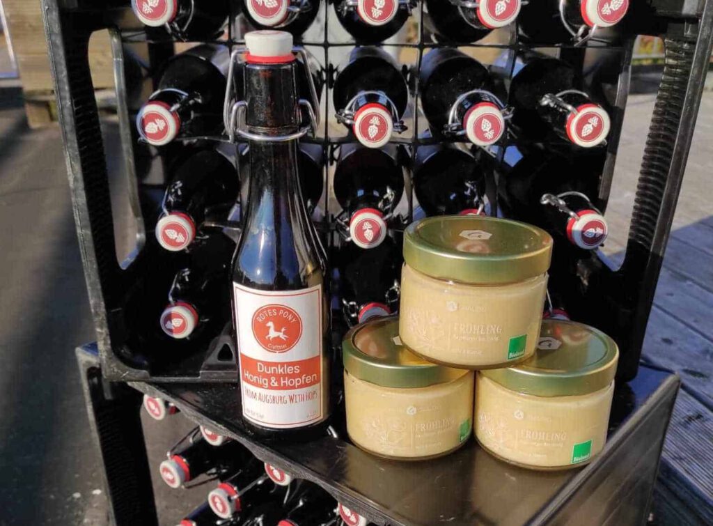 Zwei Kästen des Honig-Biers stehen übereinander. Darauf steht eine Flasche von dem Bier und 3 Gläser Frühlings-Honig der Imkerei Gräubig.
