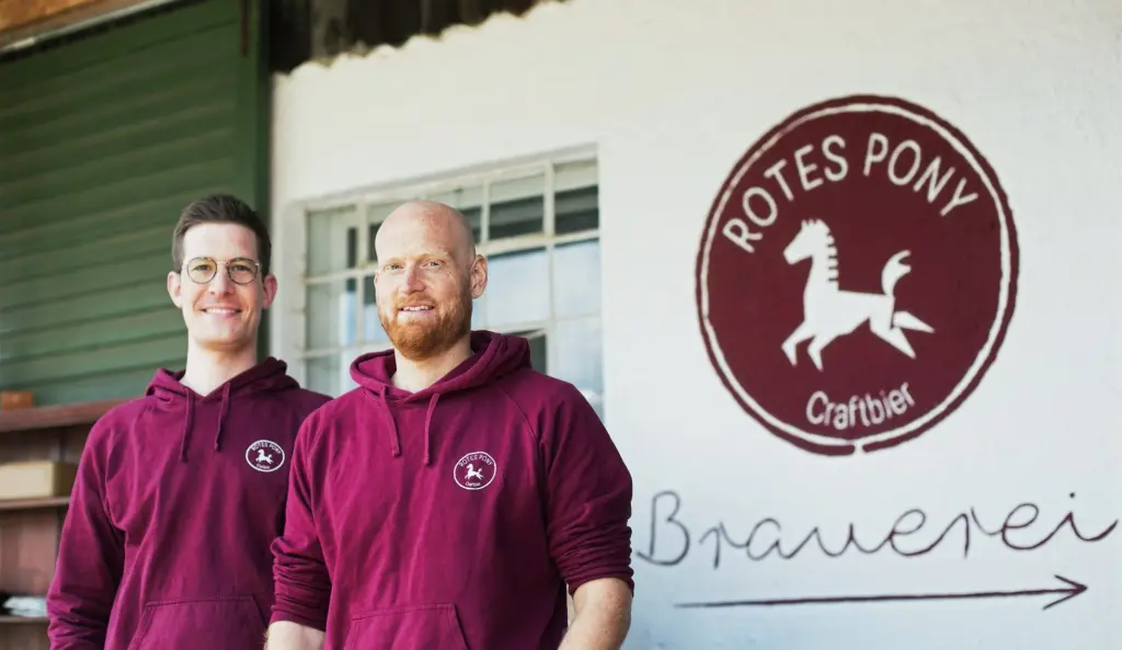 Die Gründer der Bio-Craft-Beer-Brauerei "Rotes Pony" Jerome und Christopher stehen vor ihrer Brauerei und lachen in die Kamera.